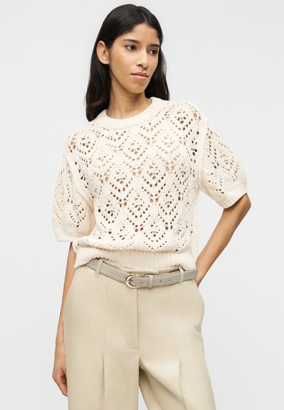 Femme portant un pull en maille ouverte crème à manches courtes, rentré dans un pantalon beige taille haute avec une ceinture gris clair, debout devant un fond uni.