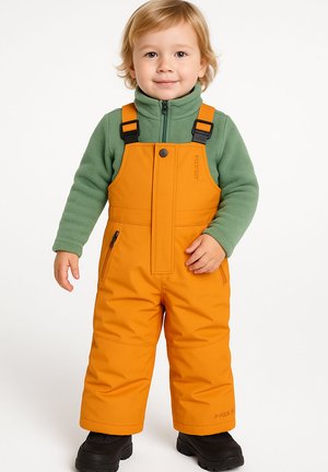 Kleinkind mit blonden Haaren, das einen grünen Fleecepullover und leuchtend orangefarbene Schneeanzug-Hosen trägt und vor einem weißen Hintergrund steht.