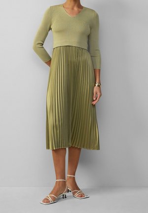Kobieta w jasnozielonej, plisowanej sukience midi z rękawami do połowy przedramienia i białych, wiązanych sandałach na obcasie na jednolitym tle.