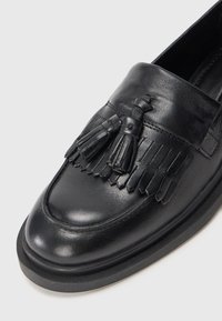 Melnas ādas loafers ar izteiksmīgiem pušķu un fringu elementiem. Apavus raksturo noapaļota purngalu forma un bieza melna zole. Gluda, spīdīga tekstūra.