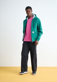 Młody mężczyzna w turkusowej bluzie na zamek, pod którą ma różową koszulkę, czarne spodnie o szerokich nogawkach i sneakersy, stoi na żółtej podłodze przed białą ścianą.