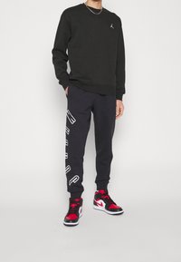 Felpa nera con un piccolo logo ricamato, abbinata a pantaloni della tuta blu navy con testo bianco e scarpe da ginnastica alte rosse e nere.