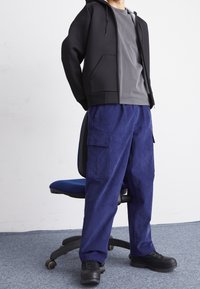 Sudadera negra con cremallera, camiseta gris y pantalones cargo azul marino con bolsillos; vestidos con zapatillas negras y gruesas; sentado en una silla de oficina azul.