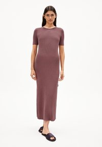 Rippent, langes kastanienbraunes Kleid mit kurzen Ärmeln und rundem Ausschnitt, im schmalen Schnitt und mit seitlichen Schlitzen. Kombiniert mit schwarzen Rutsch-Sandalen.