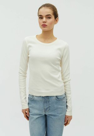 Mladá žena s vlasmi zaviazanými do drdola, oblečená v krémovom rebrovanom tričku s dlhým rukávom a svetlomodrých džínsoch, stojaca pred jednoduchým bielym pozadím.