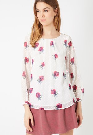 Mujer que lleva una blusa blanca de manga larga con estampados florales rojos y una falda rosa, posando frente a un fondo blanco liso.
