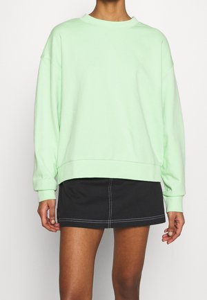 Personne portant un sweat-shirt ample vert clair et une jupe courte noire avec des coutures blanches, montrée du cou à mi-cuisse sur un fond uni.