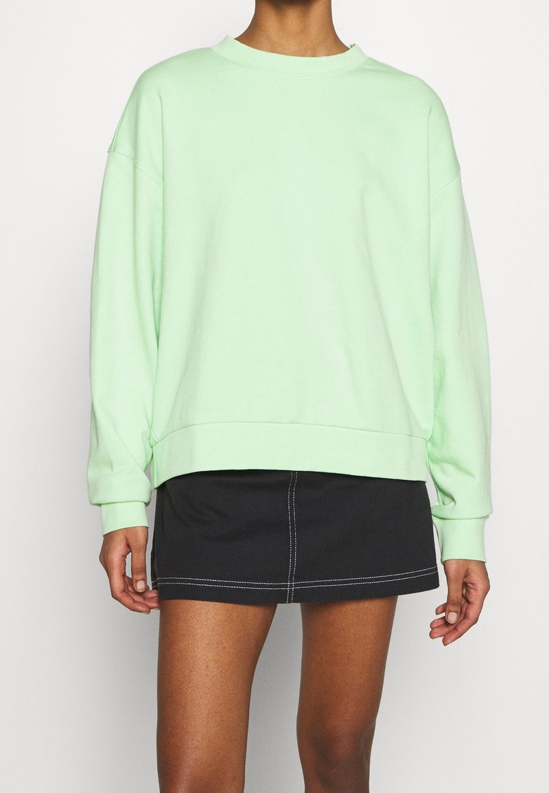 Personne portant un sweat-shirt ample vert clair et une jupe courte noire avec des coutures blanches, montrée du cou à mi-cuisse sur un fond uni.