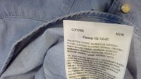 Étiquette de vêtement sur chemise bleu clair indiquant la taille, matière 100 % coton, et instructions de lavage en plusieurs langues.
