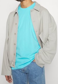 Svetle šedá oversized bunda s velkými knoflíky, nošená přes živě tyrkysové tričko. Nabízí volný střih a pohodlné džínové kalhoty.