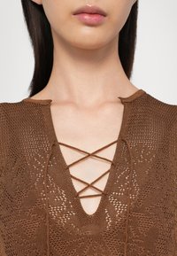 Top in maglia marrone con scollatura a V e dettagli incrociati. La texture è intricata con una finitura a motivo e cuciture visibili.
