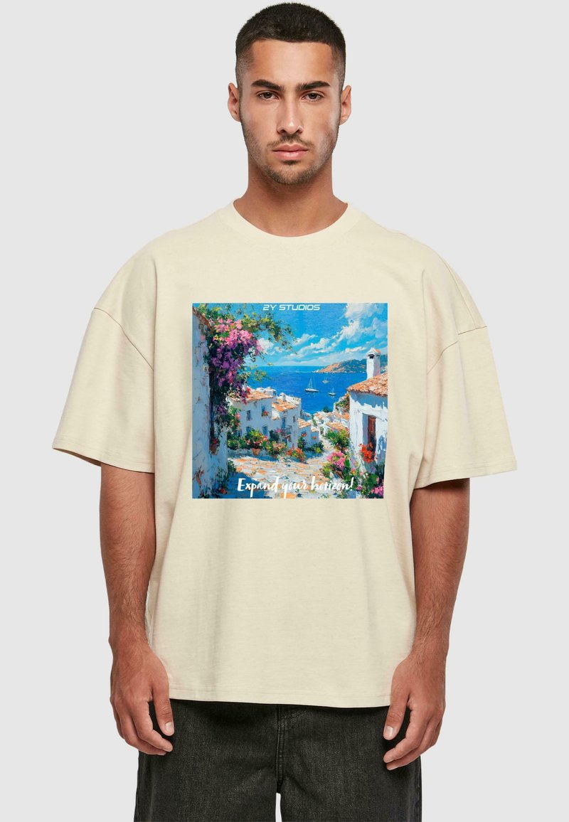 Beige per didelė t-shirt su spalvinga jūros peizažo meno spauda. Minkšta medžiaga, trumpomis rankovėmis, apvaliu kaklu ir tekstu, kuris sako "Išplėskite savo horizontus!"