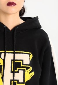 Hoodie nero con un grande design stilizzato 'F' giallo e crema con dettagli ricamati. Include un cappuccio con coulisse e una consistenza morbida.