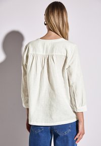 Blouse beige clair à manches trois-quarts, avec un dos froncé et des poignets boutonnés, associée à un jean bleu.