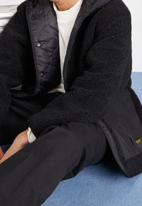 Chaqueta de forro polar negra con forro acolchado, botones a presión y bolsillos. Combinada con pantalones negros, sobre una superficie azul clara.