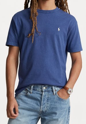 Camiseta de algodón azul con cuello redondo y mangas cortas, con un pequeño logo blanco en el pecho. Combinada con unos jeans azules descoloridos.