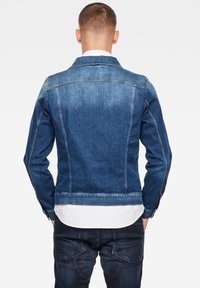 Man med blå denimjacka och mörka jeans, stående med ryggen mot kameran mot en enkel vit bakgrund.