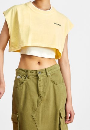 Personne portant un t-shirt Adidas court jaune sur un débardeur blanc et une jupe cargo taille haute vert olive, vue des épaules aux genoux.