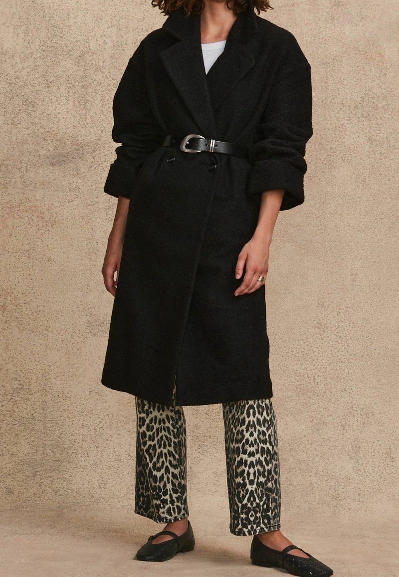 Personne portant un manteau en laine noir ceinturé, un haut blanc, un pantalon imprimé léopard et des ballerines noires, debout contre un mur beige texturé.
