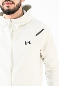 Sudadera con cremallera de color gris claro, con un cuello alto, de textura suave, con un logo negro en el pecho y un acento oscuro en el hombro.