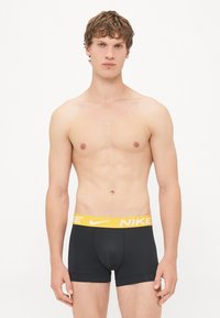 Crne bokserice s žutim pojasom Nike loga. Tkanina izgleda glatko s uskim izgledom i minimalnim šavovima.