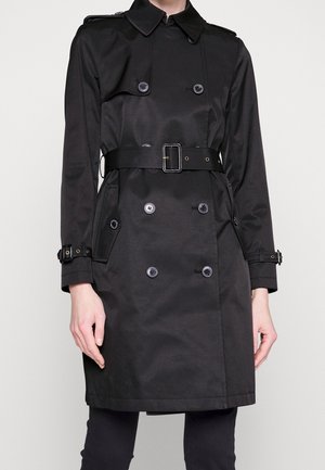 Černý trenčkot s páskem, dvouřadými knoflíky a přezkami na rukávech, nošený osobou na jednobarevném pozadí.