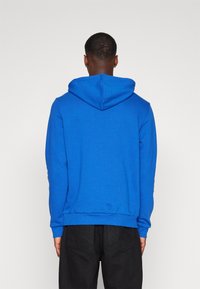 Blå bomullshoodie med dragsko i huvan, ribbade muddar och midjeband; slät yta, avslappnad design. Svarta byxor syns delvis.