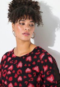 Camiseta negra con estampado de leopardo en forma de corazón rojo y rosa; cuenta con cuello redondo y detalles transparentes en las mangas. Pendientes de aro plateados visibles.