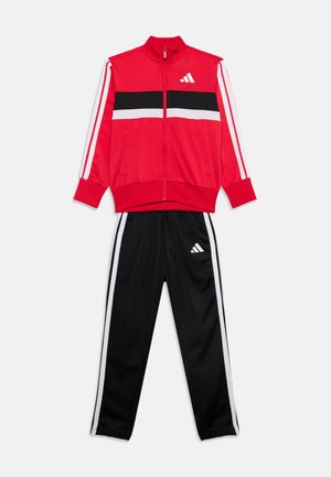 Rode sportjas met rits met zwarte en witte strepen, gecombineerd met zwarte trainingsbroek met witte zijstrepen en logo's.
