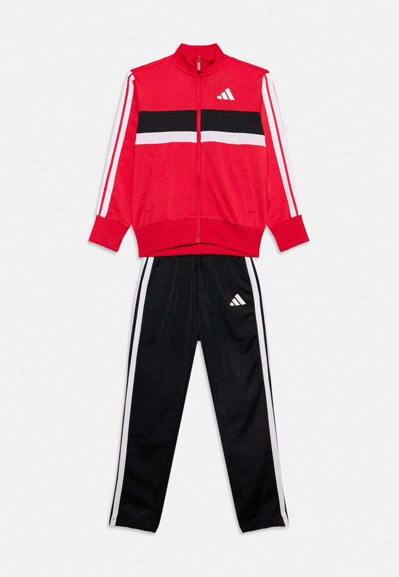 Veste de sport rouge zippée avec des rayures noires et blanches, associée à un pantalon de survêtement noir avec des bandes blanches latérales et des logos.