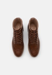 Bottines en cuir marron avec des détails de brogue décoratifs, bout rond et fermeture à lacets. Texture lisse avec un motif de couture distinct.