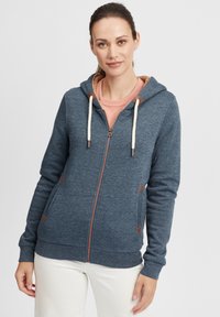 Sweat à capuche zippé en tissu bleu foncé, avec des accents beige, des cordons de serrage et des poches latérales avec des détails triangulaires.