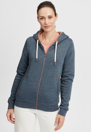 Sweat à capuche zippé en tissu bleu foncé, avec des accents beige, des cordons de serrage et des poches latérales avec des détails triangulaires.