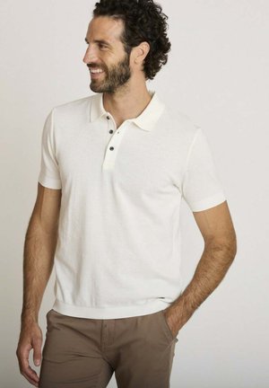 Homme souriant et regardant vers la gauche, portant un polo blanc à manches courtes et un pantalon marron, une main dans la poche.