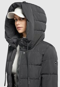 Giacca imbottita di un grigio scuro con un ampio cappuccio. Presenta una chiusura frontale con zip, lacci e dettagli con bottoni sulle maniche.