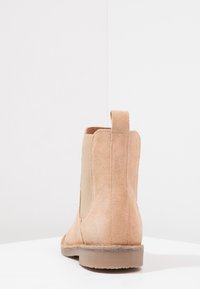 Botín de ante marrón con paneles laterales elásticos, tirador y suela de goma plana. Presenta un diseño limpio y estilizado con una textura suave.