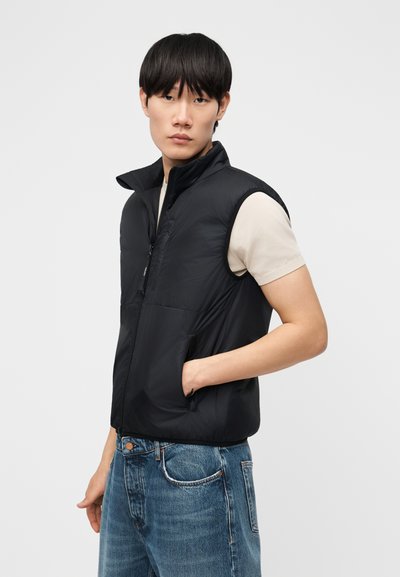 Νέος άνδρας με μαύρα μαλλιά, φορώντας μαύρο αμάνικο γιλέκο με φερμουάρ, μπεζ κοντομάνικο πουκάμισο και μπλε τζιν, ποζάρει με το χέρι στην τσέπη.