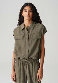 Olijfgroene mouwloze shirt met een knoopsluiting aan de voorkant, twee borstzakken en een gerimpelde taille met trekkoord. Lichtgewicht, gestructureerde stof.