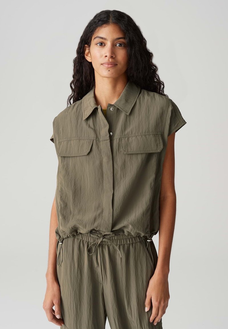 Olijfgroene mouwloze shirt met een knoopsluiting aan de voorkant, twee borstzakken en een gerimpelde taille met trekkoord. Lichtgewicht, gestructureerde stof.