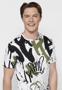 Camiseta blanca con un diseño inspirado en graffiti, salpicaduras en negro y verde olivo. Mangas cortas y cuello redondo.