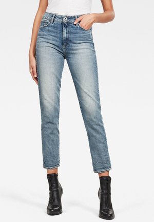 Kvinde iført lyseblå højtaljede cropped jeans, hvid top og sorte ankellange støvler med lynlåse foran, stående med den ene hånd i lommen.