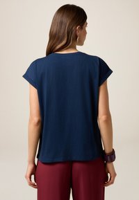 Tricou din bumbac bleumarin, cu mâneci scurte rulate, guler ribbat, margine rotundă, purtat cu pantaloni vișinii și o brățară mov.