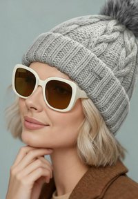Gray knitted hat with a pom-pom, featuring a braided design. Large white sunglasses with brown lenses, worn resting on the cheek.