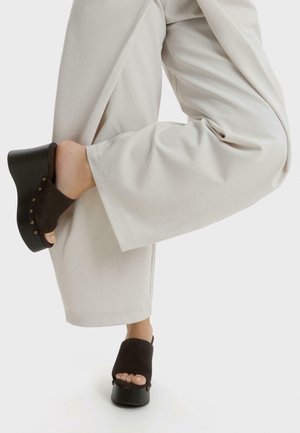 Persoon die beige wijde broek en zwarte platform klompen met open teen draagt, tilt één knie gebogen been op tegen een witte achtergrond.