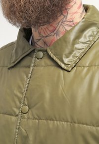 Olivgrüne Pufferjacke mit einer glatten, glänzenden Textur, ausgestattet mit einem umklappbaren Kragen und Druckknopfverschluss.