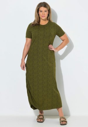 Frau steht mit der Hand in der Hüfte, trägt ein olivgrünes kurzärmeliges Strick-Maxikleid und passende Sandalen mit Schnalle vor weißem Hintergrund.