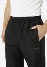 Schwarze Sporthosen mit elastischem Bund, Seitentaschen und einem auf der linken Seite aufgedruckten Logo. Glatte, leichte Stofftextur.