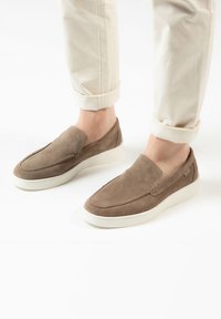 Brown suede slip-on shoes with a rounded toe, white rubber sole, and minimal stitching details. Worn with rolled cream trousers.