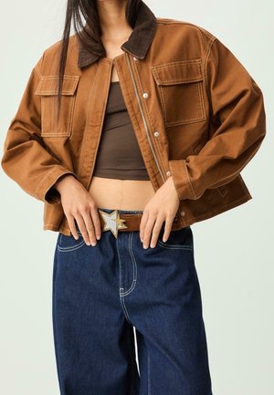Vrouw draagt een kort bruin jasje over een donkerbruin cropped top, losse donkerblauwe jeans en een riem met een stervormige gesp, poseert met handen bij de taille.