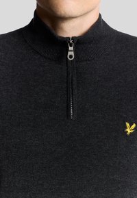 Svart zip-necksweater gjord av stickad tyg. Har en ribbad krage och en gul broderad örnlogotyp på vänster sida.